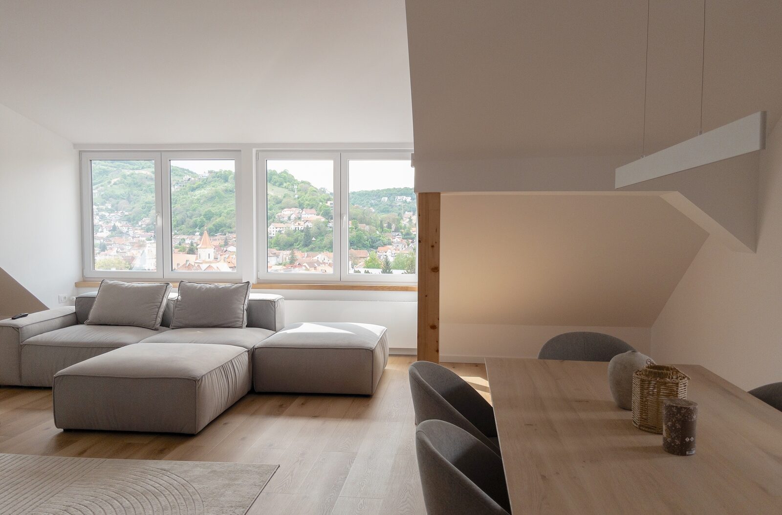 The finished flat: a pale sofa and an oak dining table under the roof slope, with a long band of windows looking over a hillside town.